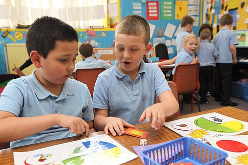 Two children sitting at a desk playing a game, another looks over from afar.